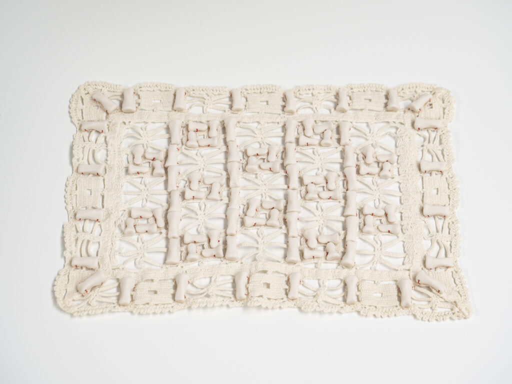An image of Blake's piece, titled Threads (Anagnorisis). White porcelain finger bones sewn onto a rectangular vintage textile with red thread.